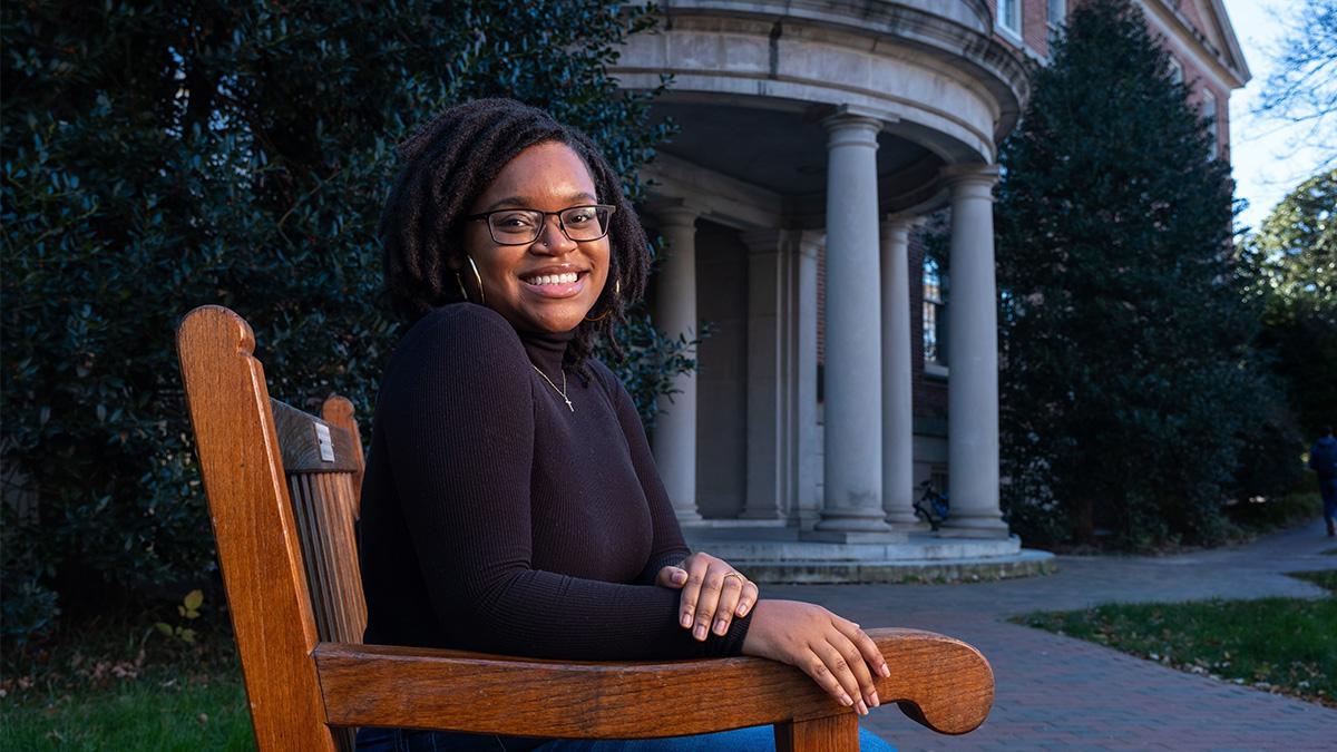 Dezja Ishmeal sitting on a bench on U.N.C. campus with the Old Well in the background.