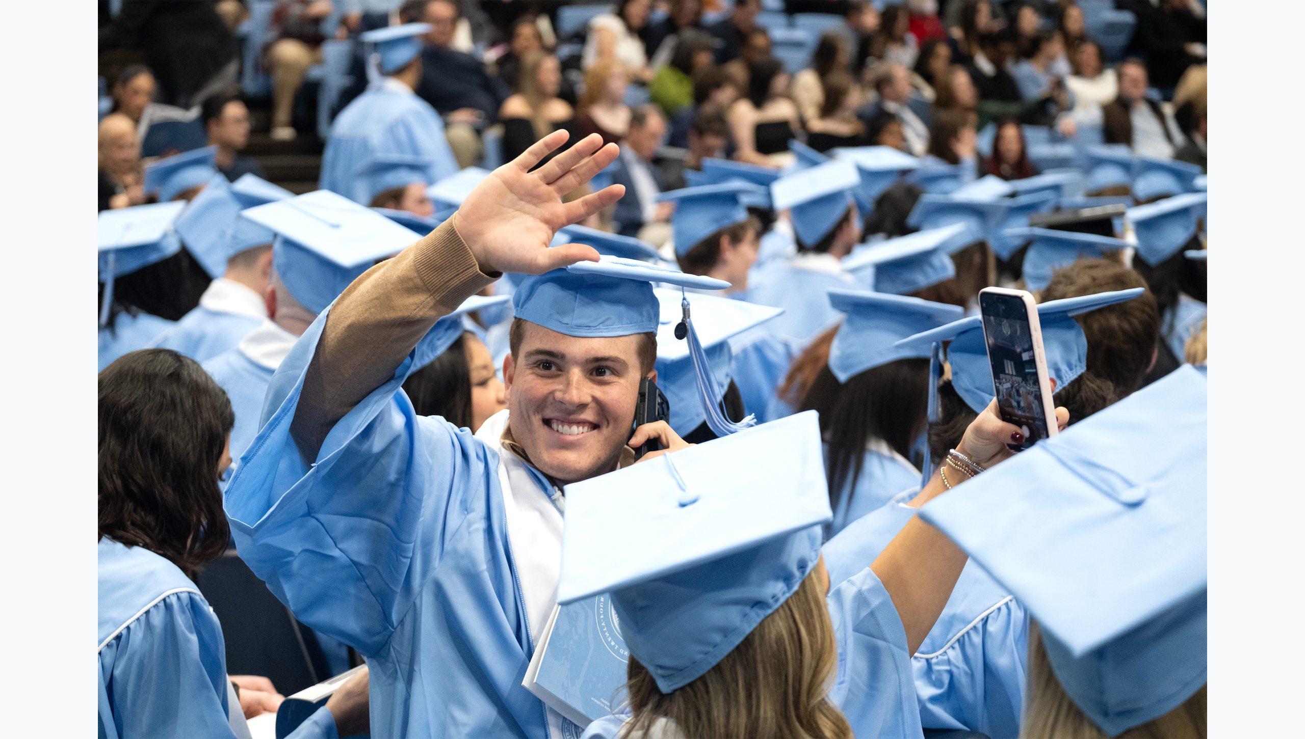 A male student at U.N.C. Chapel Hill Winter Commencement on the phone and waving to someone.